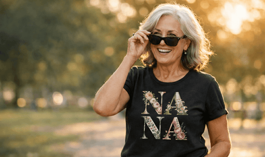 A smiling older woman with grey hair wearing sunglasses and a black t-shirt featuring a floral "NANA" typography design, standing outdoors in a soft-focus park setting at sunset.