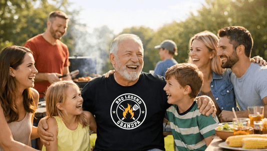 Grandfather in a “BBQ Legend Grandpa” t‑shirt smiling and sitting at an outdoor picnic table, surrounded by his children and grandchildren at a sunny backyard barbecue.