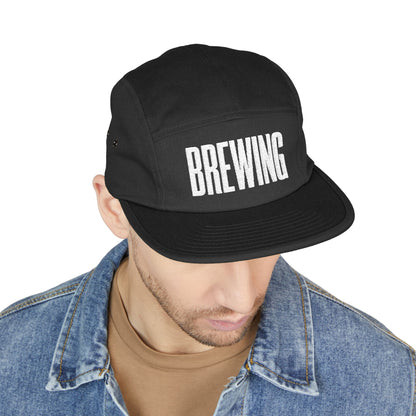 Black cap with 'BREWING' text worn by a person on a white background. BREWING minimalist text embroidery design on 5-panel cap. TEEZOCA