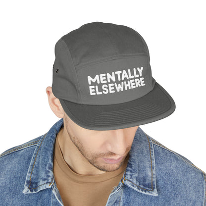 Person wearing a gray cap with 'MENTALLY ELSEWHERE' text on a white background. Mentally Elsewhere embroidered text design on five-panel cap. TEEZOCA