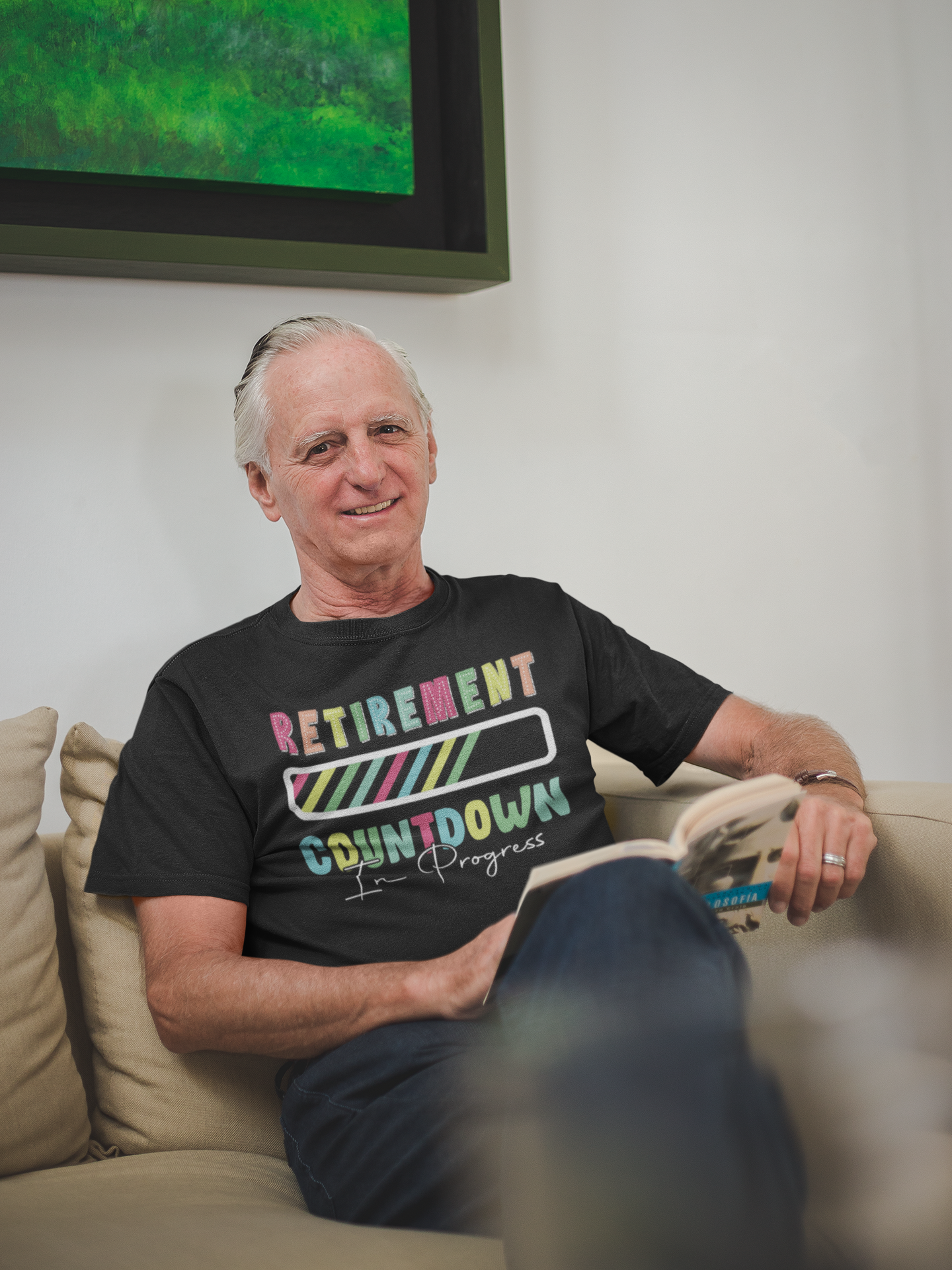 “Older man sitting on couch reading a book while wearing a black Teezoca Retirement Countdown In Progress t-shirt, lifestyle mockup, front view.”
