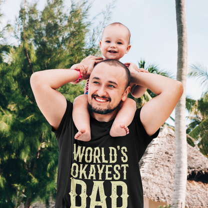  Smiling dad wearing “WORLD’S OKAYEST DAD” T‑shirt with child on shoulders outdoors, lifestyle mockup photo.