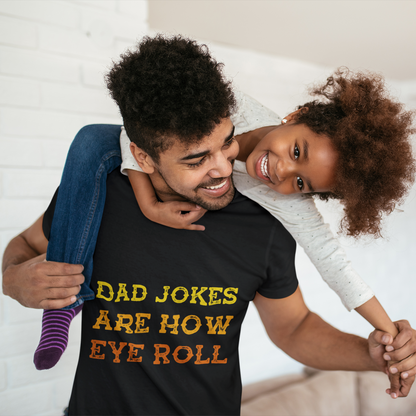 Man wearing a TEEZOCA black t-shirt with 'Dad jokes are how eye roll' text, holding a child in a living room