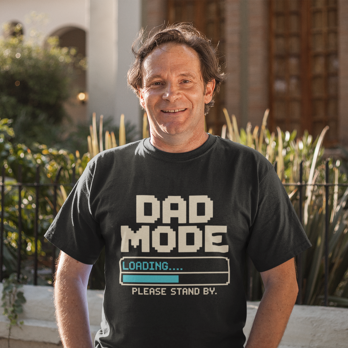 Smiling man wearing a TEEZOCA black “Dad Mode Loading Please Stand By” funny gamer dad t‑shirt outdoors.
