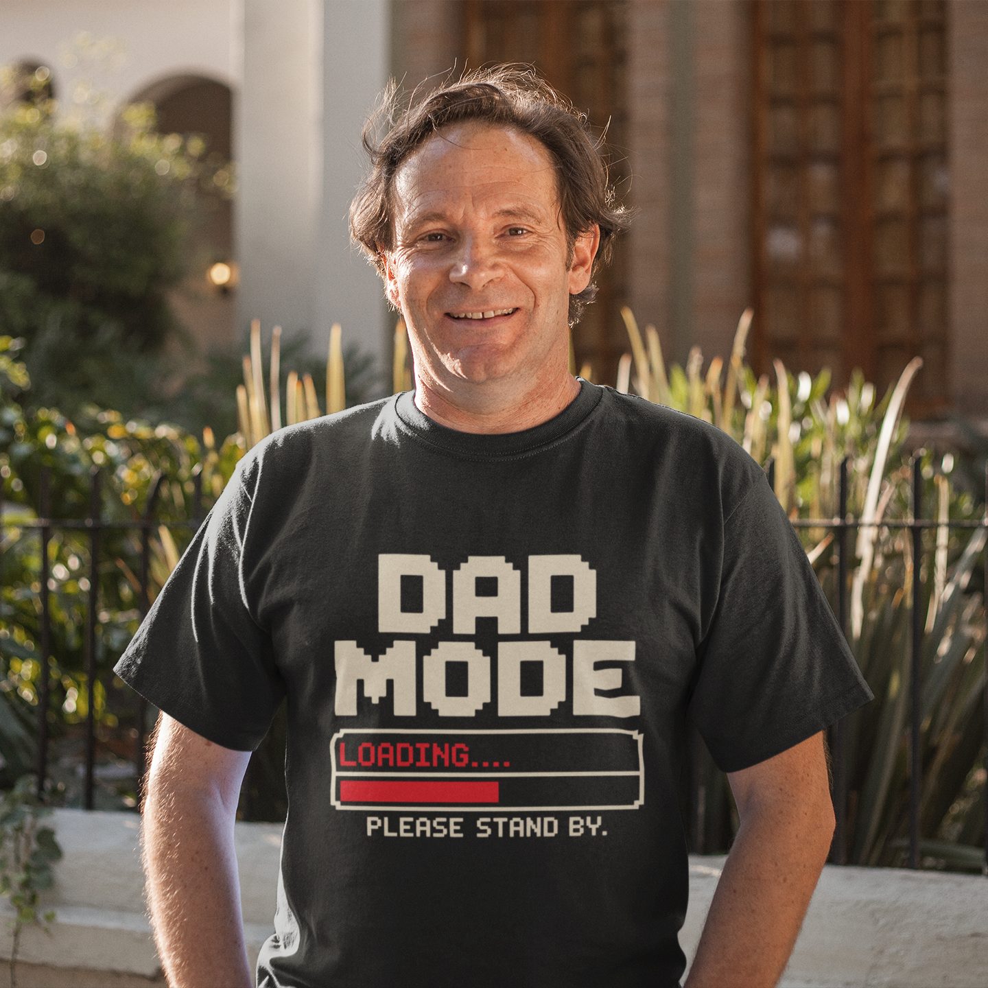 Smiling man wearing a TEEZOCA “Dad Mode Loading Please Stand By” funny dad t-shirt outdoors.
