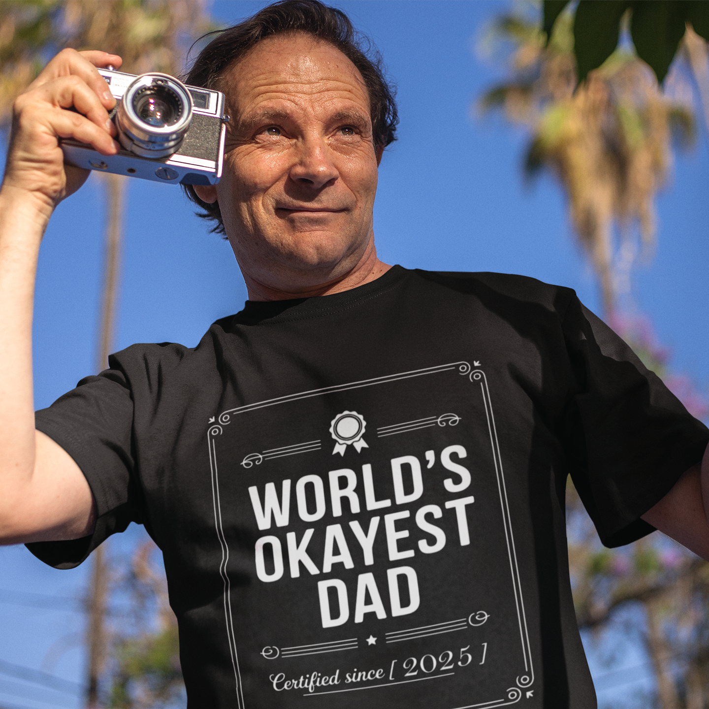 Smiling man holding up a TEEZOCA “World’s Okayest Dad Certified Since 2025” funny t-shirt outdoors.