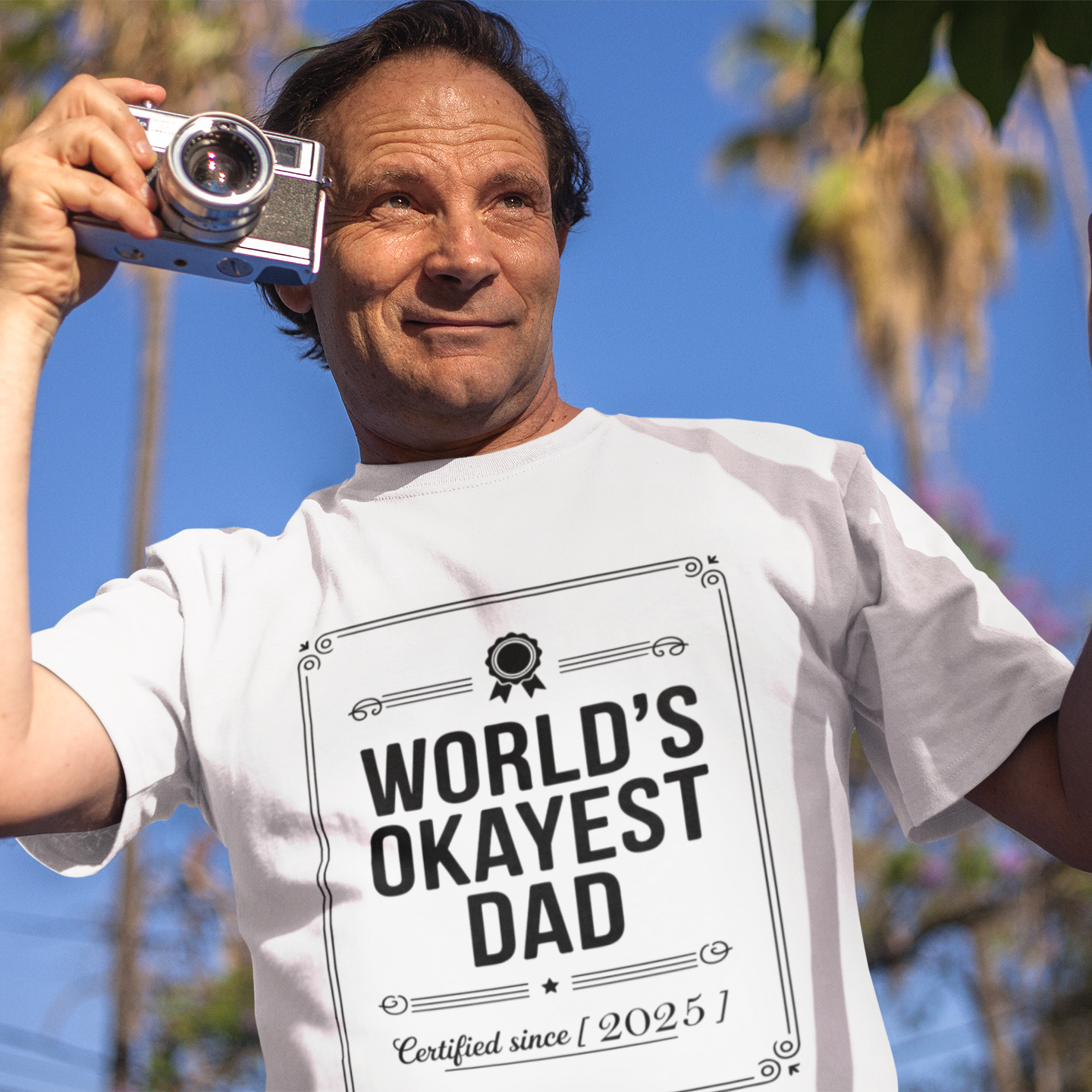 Smiling man wearing a white TEEZOCA “World’s Okayest Dad Certified Since 2025” graphic t-shirt outdoors.