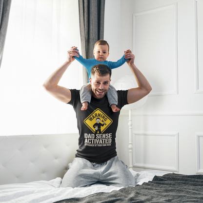 Man lifting a child while wearing a black TEEZOCA “Warning Dad Sense Activated” graphic t-shirt.
