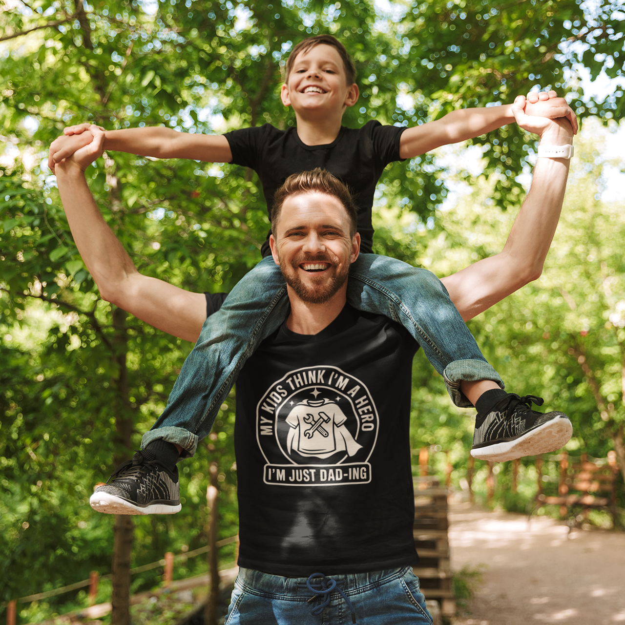 Dad wearing a TEEZOCA “My Kids Think I’m A Hero I’m Just Dad‑ing” T‑shirt while carrying a child outdoors.