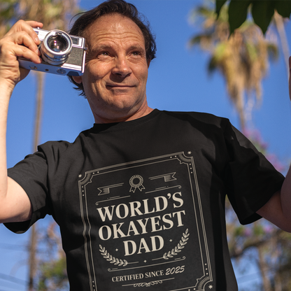 Smiling man wearing a black TEEZOCA “World’s Okayest Dad Certified Since 2025” funny dad T‑shirt outdoors.