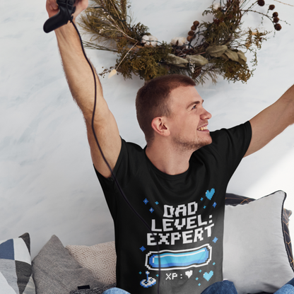 Man cheering on a couch while wearing a black TEEZOCA “Dad Level: Expert” gamer dad T‑shirt.