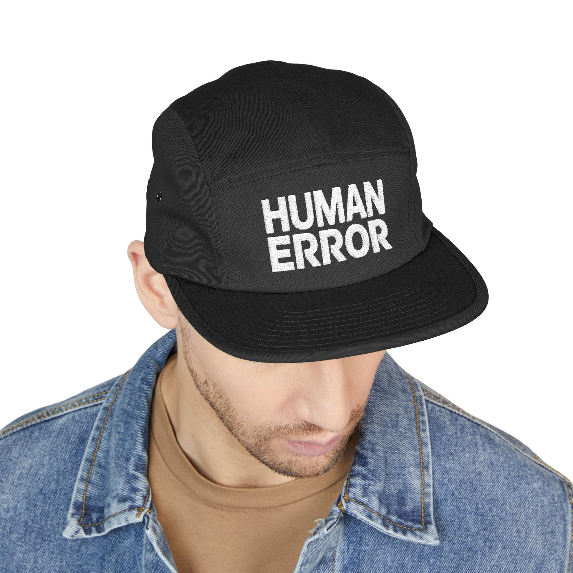 Black cap with 'HUMAN ERROR' text worn by a person on a white background. Lifestyle shot of a model wearing the HUMAN ERROR embroidered cap in a casual streetwear outfit.