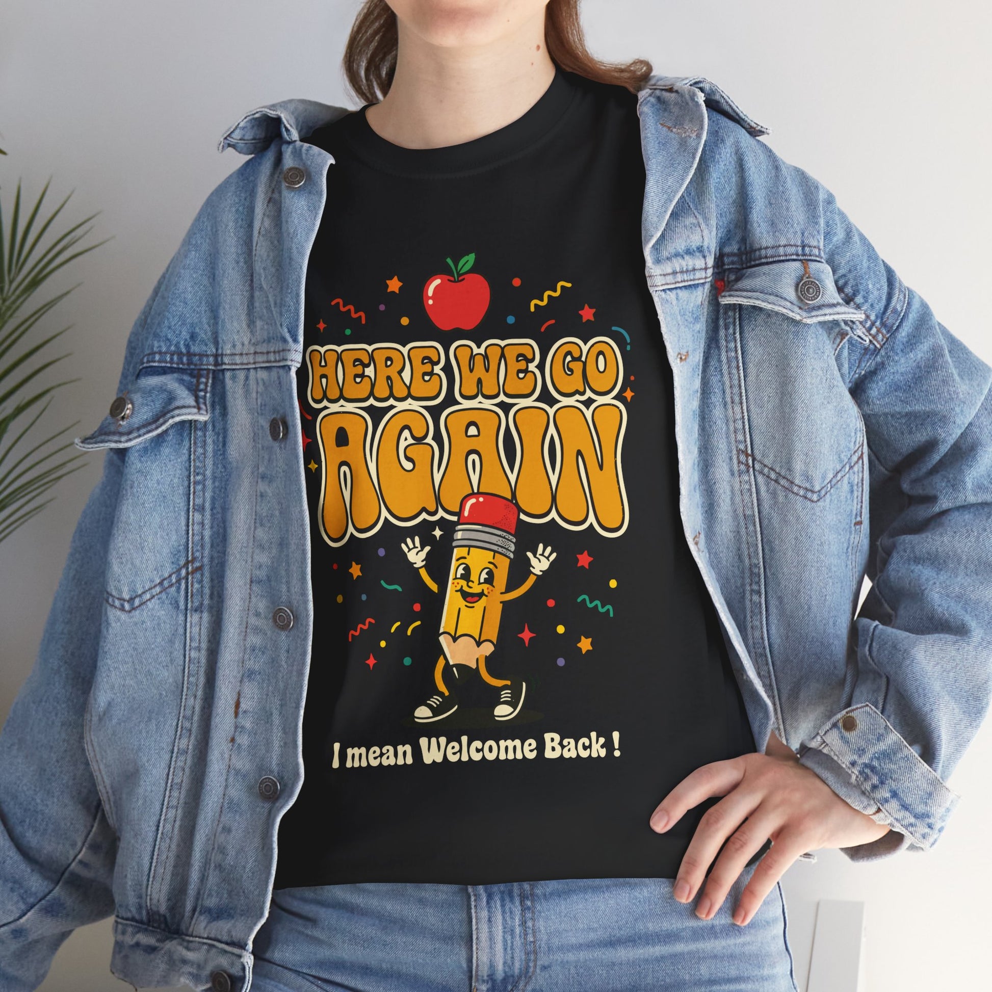 Black T-shirt featuring a funny back to school design with the words “Here We Go Again” and “I mean Welcome Back!” plus a red apple and a pencil character.
