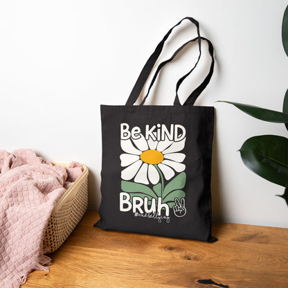 “Black canvas tote bag with white daisy and ‘Be Kind Bruh’ message resting on wooden table beside pink throw and green plant, front view”


