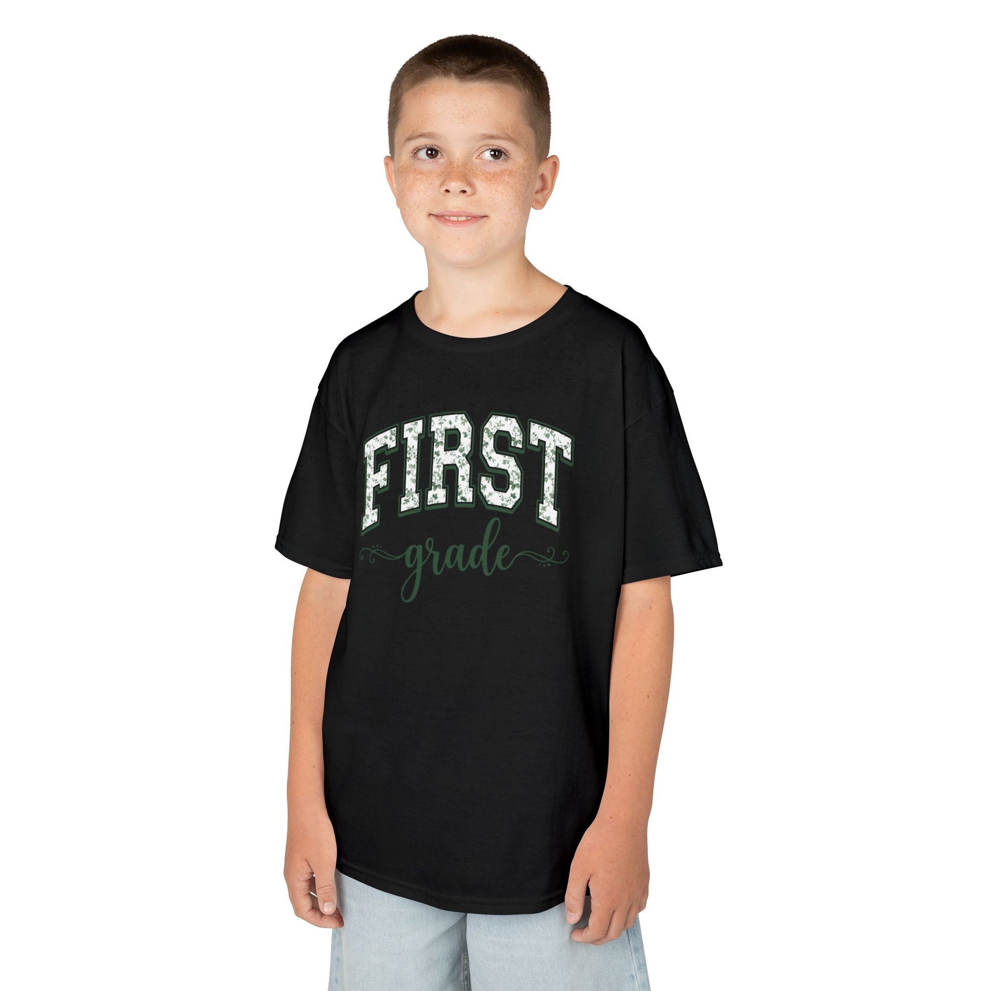Child wearing a black t-shirt with 'FIRST grade' text on a white background. Botanical first grade shirt for kids by TEEZOCA with hand-lettered script and a soft eucalyptus-green palette.
