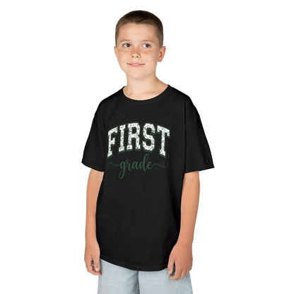 Child wearing a black t-shirt with 'FIRST grade' text on a white background. Botanical first grade shirt for kids by TEEZOCA with hand-lettered script and a soft eucalyptus-green palette.

