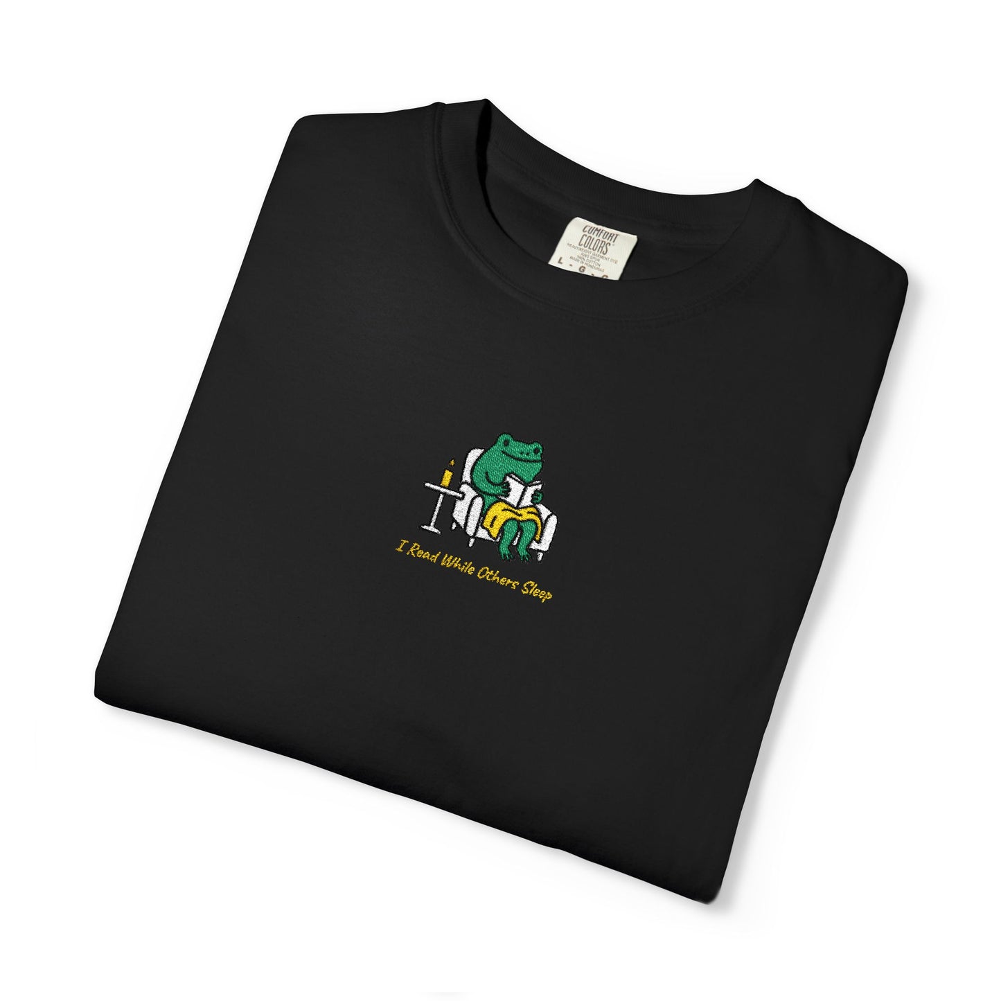  Close-up of a Black Comfort Colors® shirt featuring a cozy green frog reading in an armchair by candlelight with the saying “I Read While Others Sleep.