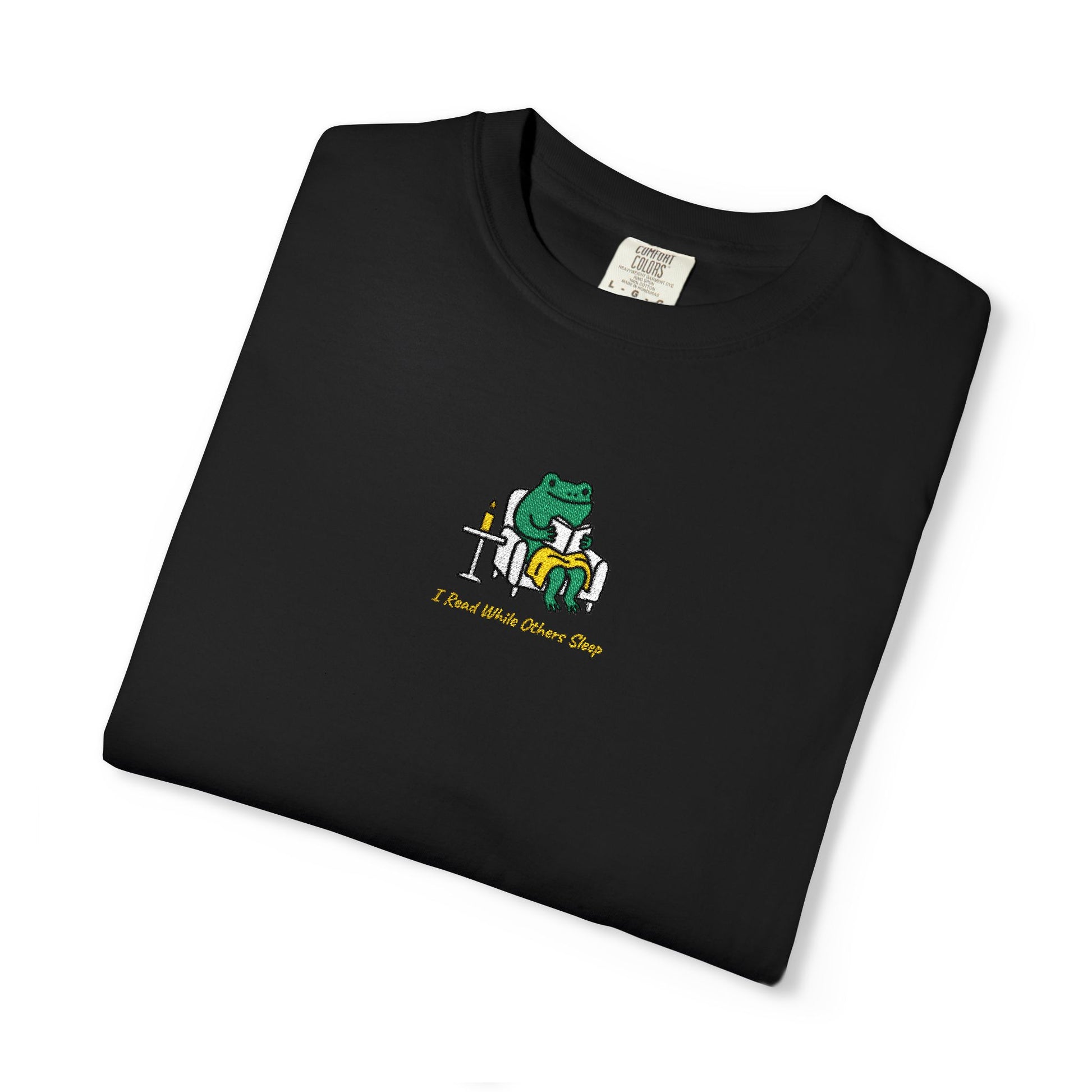  Close-up of a Black Comfort Colors® shirt featuring a cozy green frog reading in an armchair by candlelight with the saying “I Read While Others Sleep.