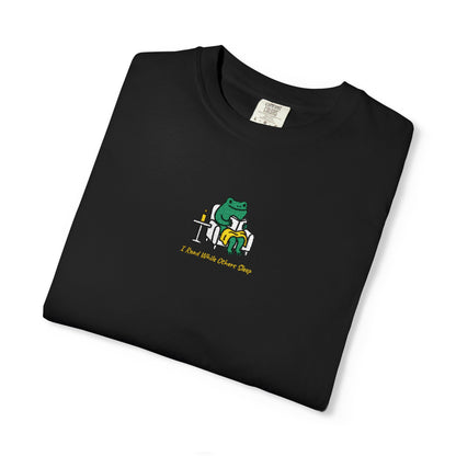  Close-up of a Black Comfort Colors® shirt featuring a cozy green frog reading in an armchair by candlelight with the saying “I Read While Others Sleep.