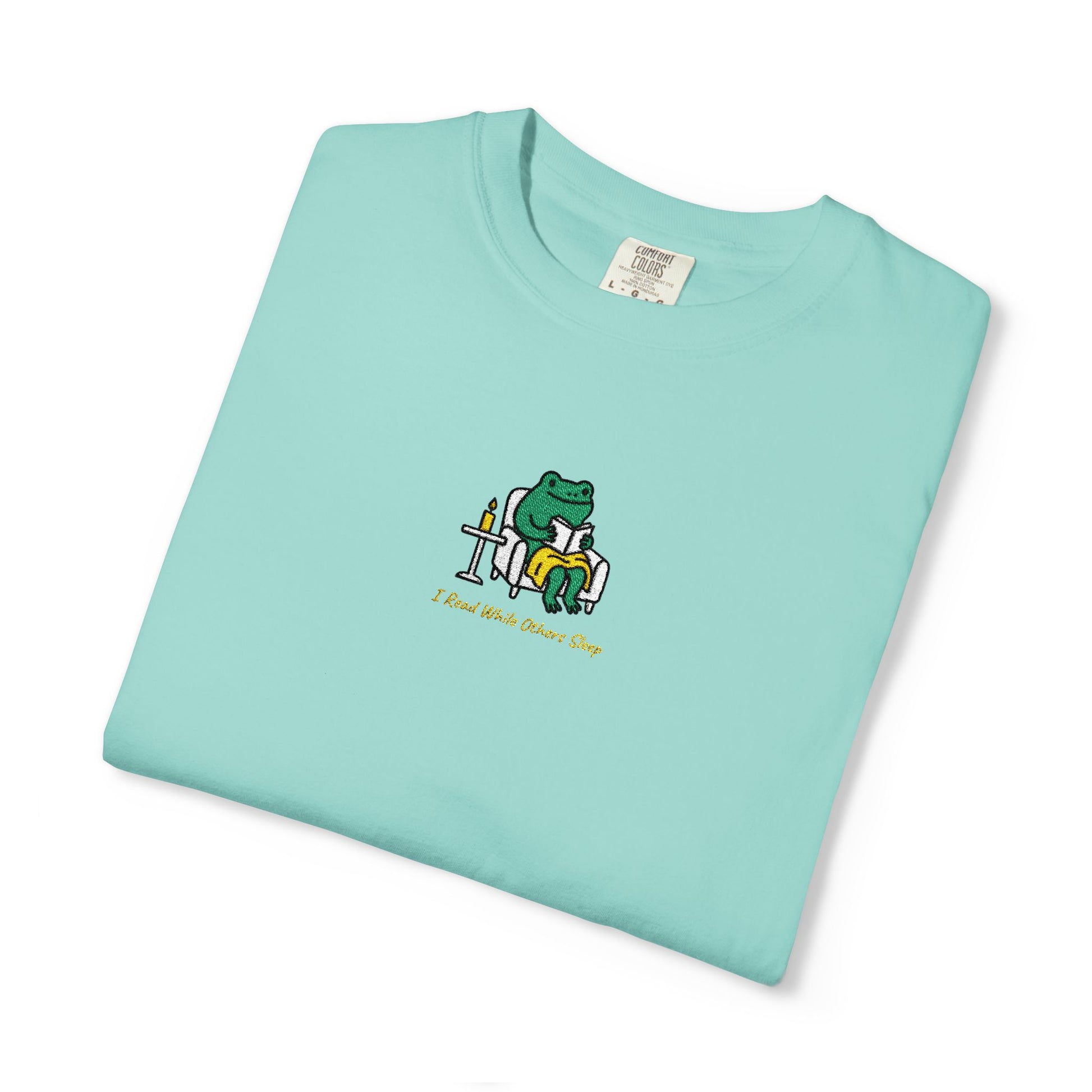  Close-up of a Chalky Mint Comfort Colors® shirt featuring a cozy green frog reading in an armchair by candlelight with the saying “I Read While Others Sleep.
