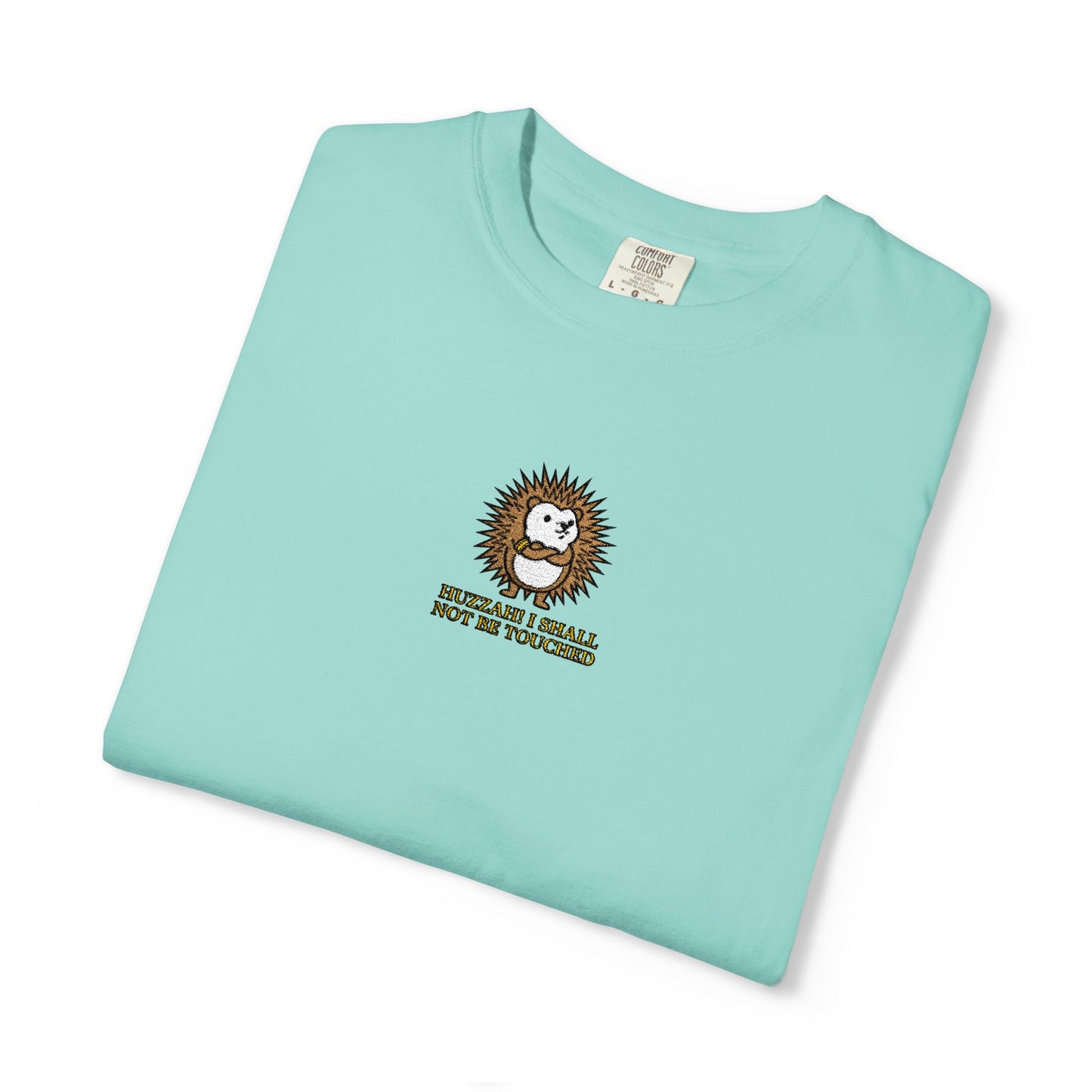 Close-up of a Chalky Mint Comfort Colors® shirt featuring an embroidered round hedgehog with gold lettering saying "Huzzah! I Shall Not Be Touched."

