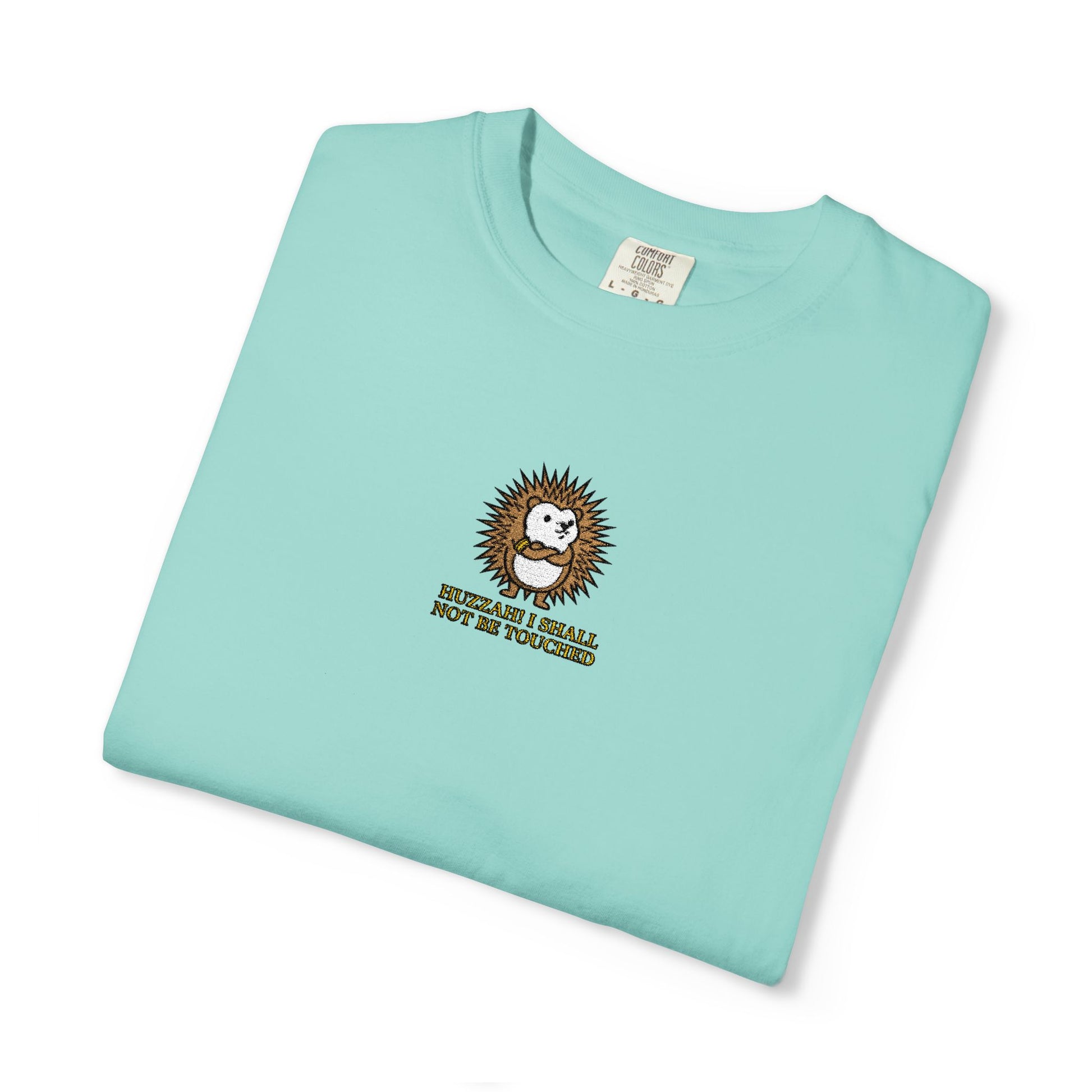Close-up of a Chalky Mint Comfort Colors® shirt featuring an embroidered round hedgehog with gold lettering saying "Huzzah! I Shall Not Be Touched."

