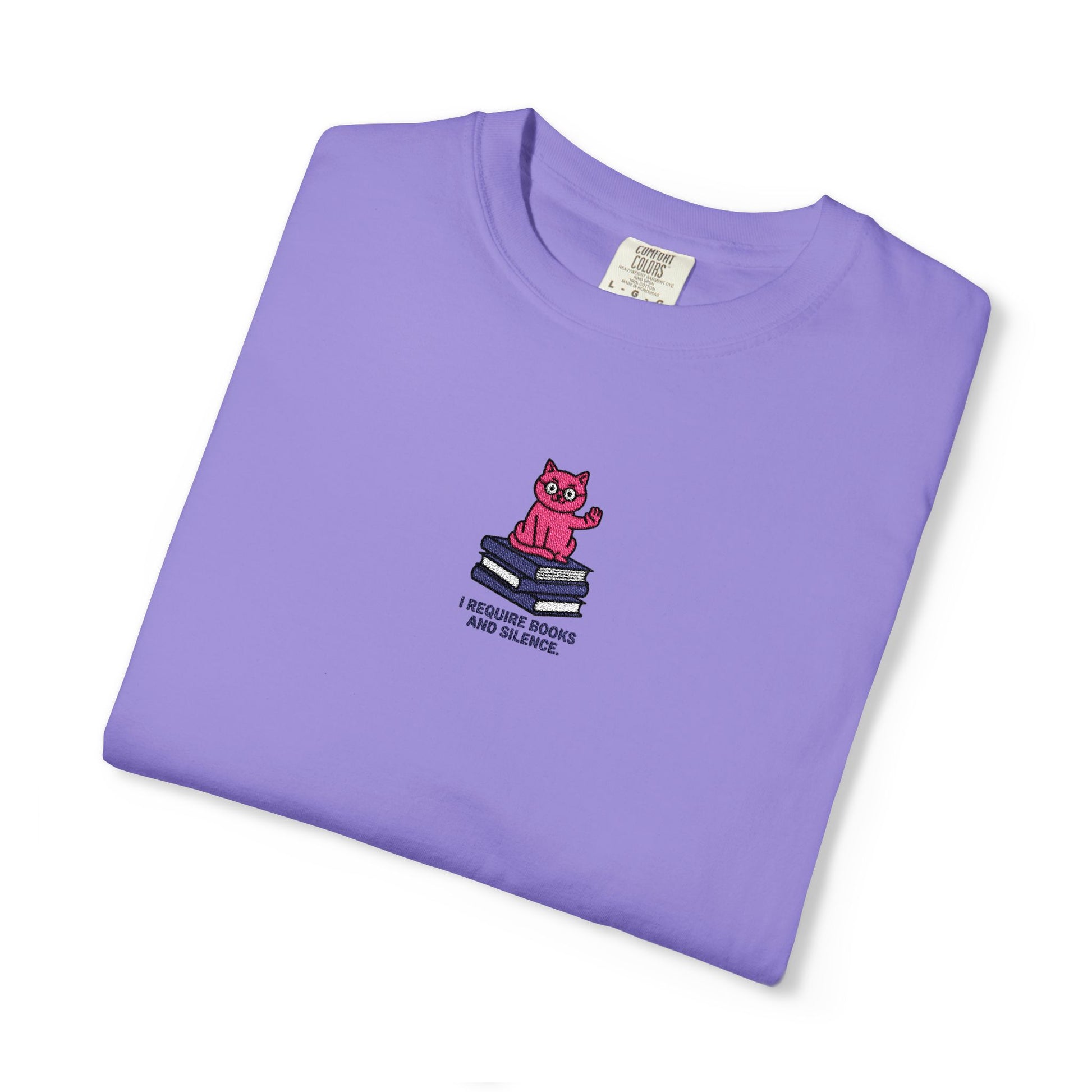   Close-up of a Comfort Colors®  Voilet shirt featuring an embroidered pink cat sitting on books with the saying “I Require Books and Silence.”

