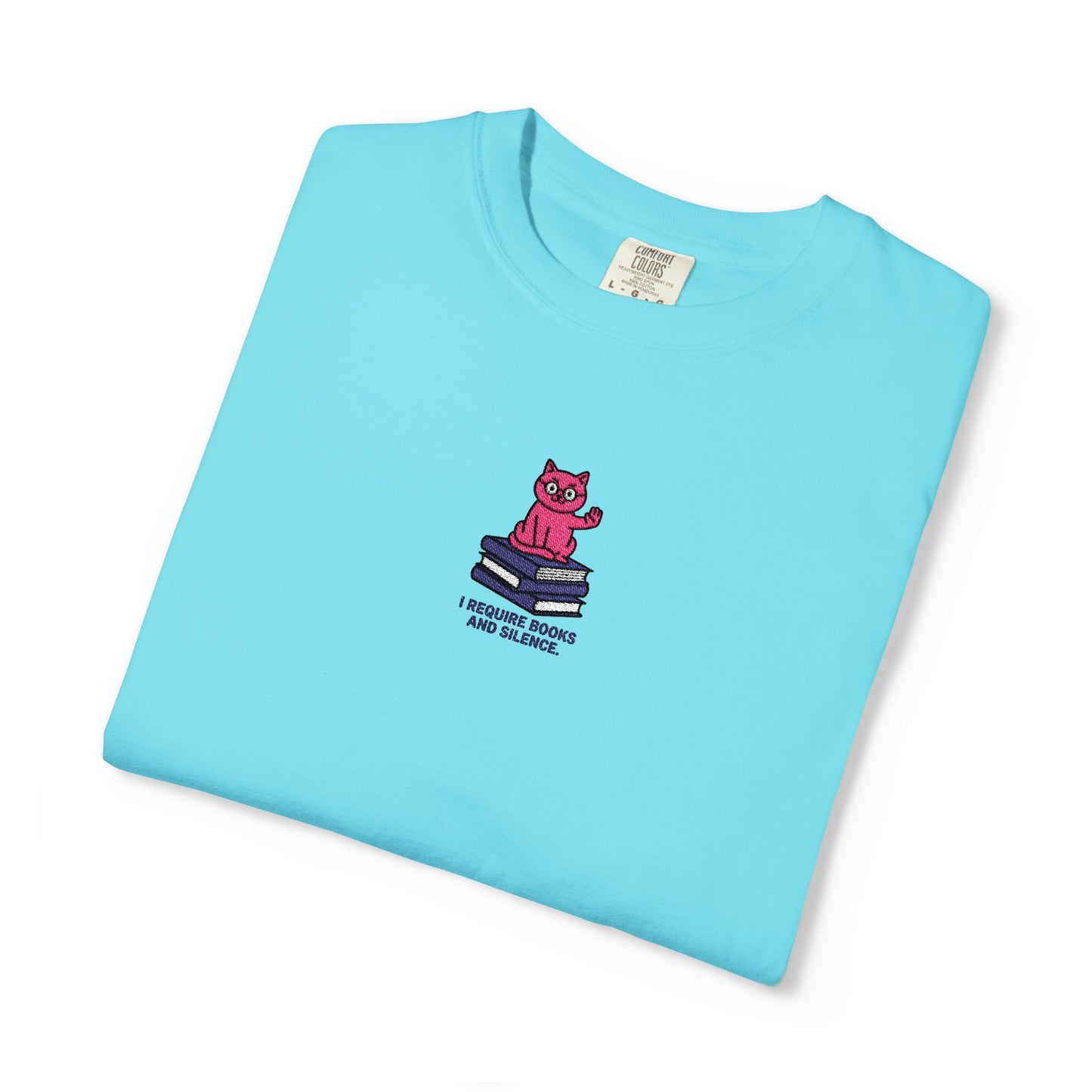 Close-up of a Comfort Colors®  lagoon blue shirt featuring an embroidered pink cat sitting on books with the saying “I Require Books and Silence.”

