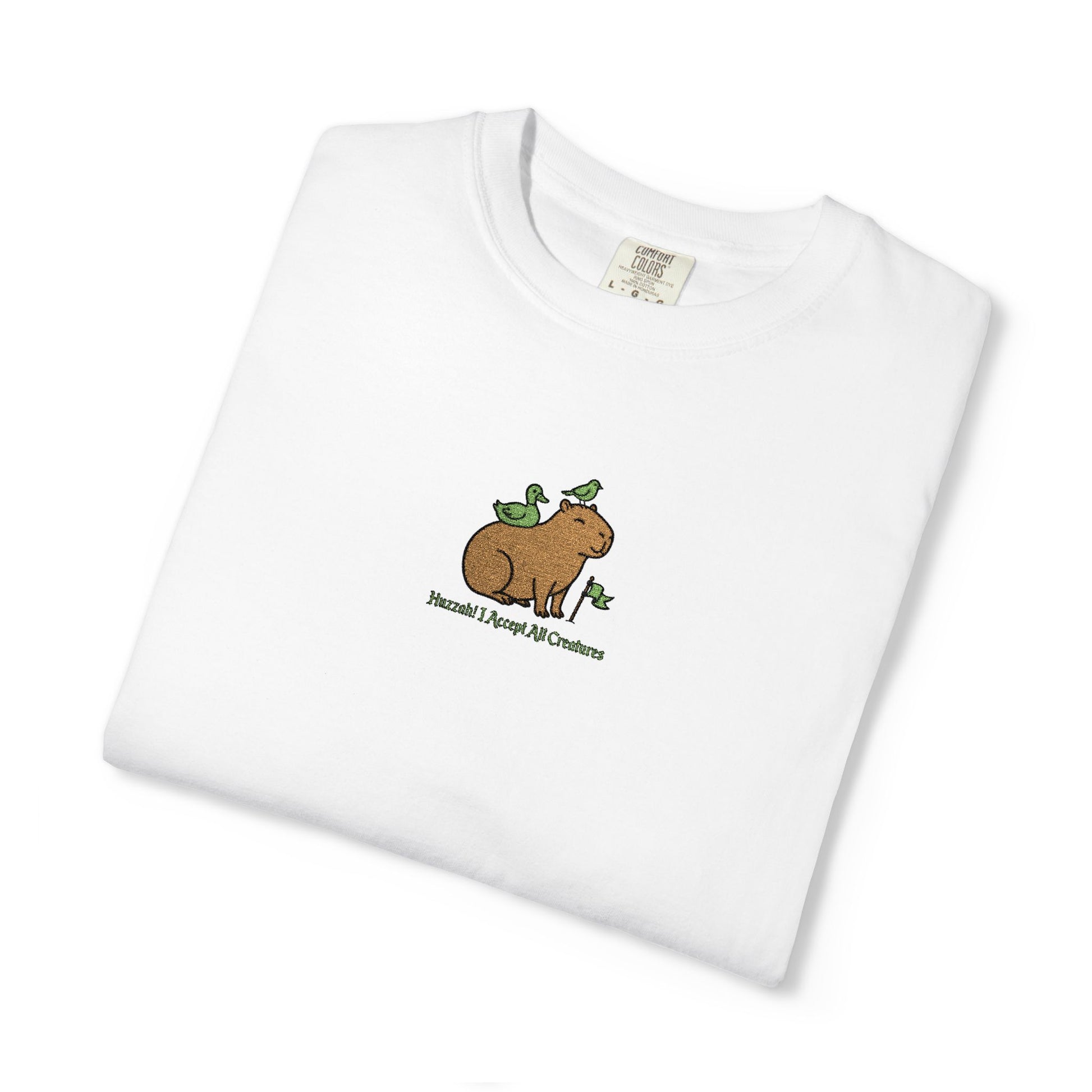 Close-up of a Comfort Colors® white shirt featuring an embroidered brown capybara with two green birds and the saying “Huzzah! I Accept All Creatures.”
