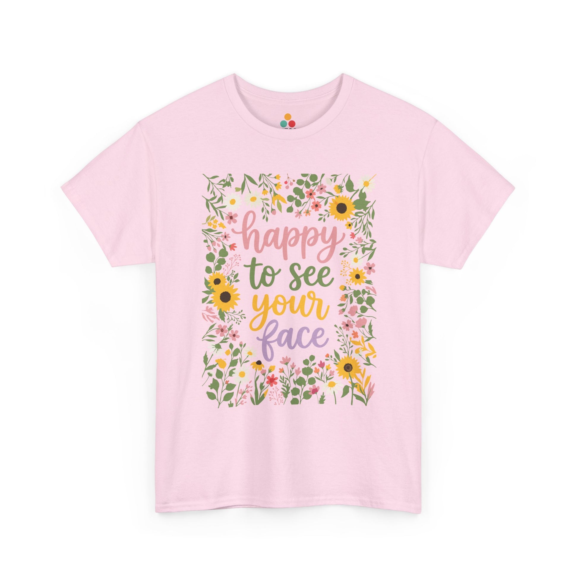 Close-up of a PinkT-shirt featuring the phrase “Happy to See Your Face” in pastel lettering framed by a floral border with sunflowers and wildflowers.

