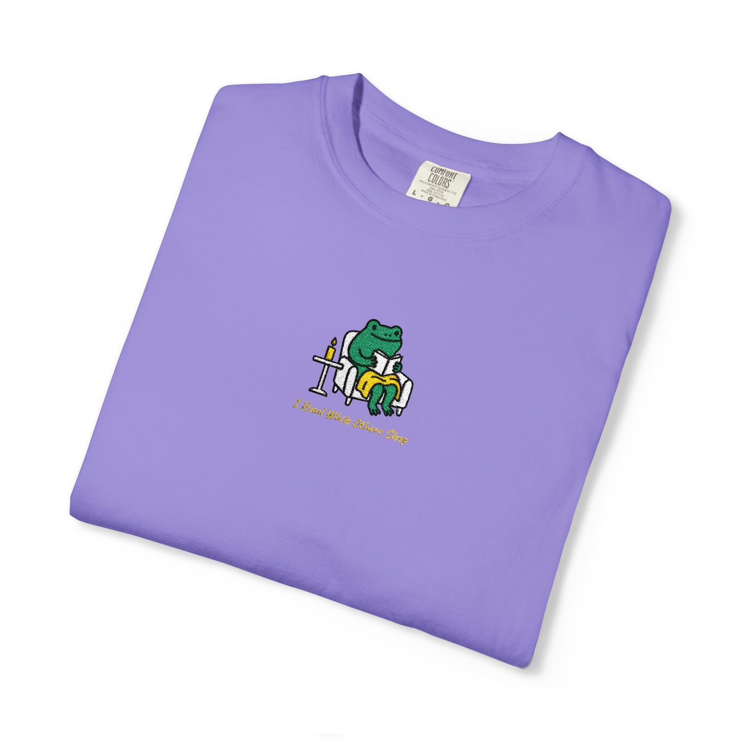  Close-up of a Violet Comfort Colors® shirt featuring a cozy green frog reading in an armchair by candlelight with the saying “I Read While Others Sleep.