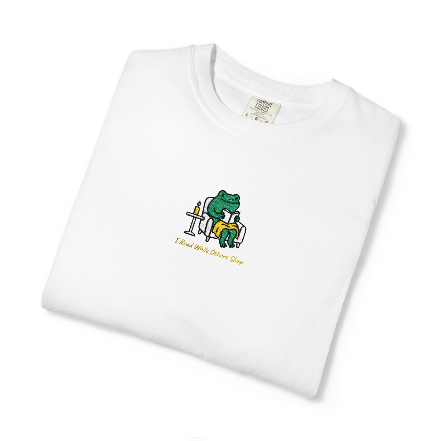 Close-up of a White Comfort Colors® shirt featuring a cozy green frog reading in an armchair by candlelight with the saying “I Read While Others Sleep.