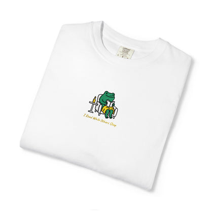 Close-up of a White Comfort Colors® shirt featuring a cozy green frog reading in an armchair by candlelight with the saying “I Read While Others Sleep.