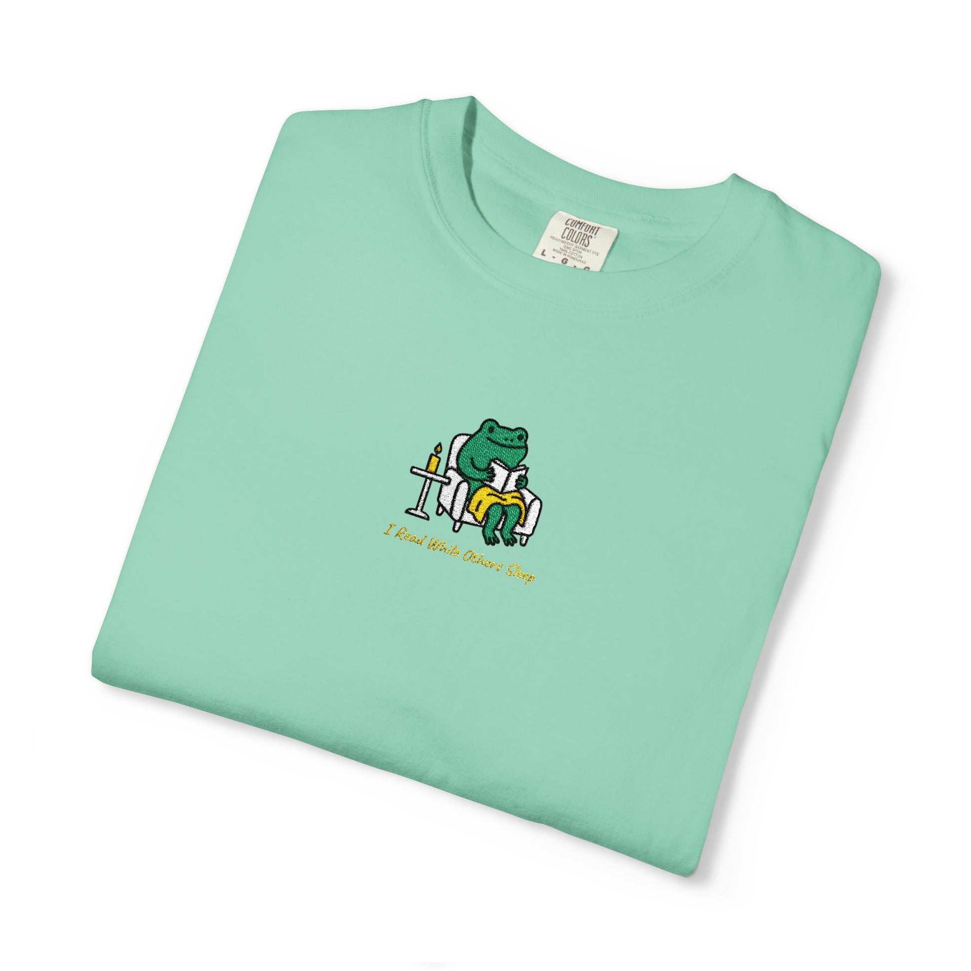  Close-up of a island reef Comfort Colors® shirt featuring a cozy green frog reading in an armchair by candlelight with the saying “I Read While Others Sleep.