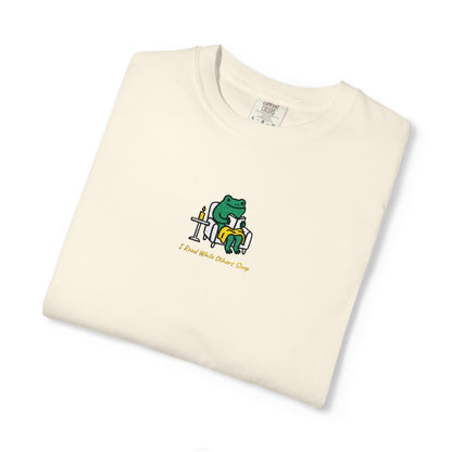 Close-up of a ivory Comfort Colors® shirt featuring a cozy green frog reading in an armchair by candlelight with the saying “I Read While Others Sleep.