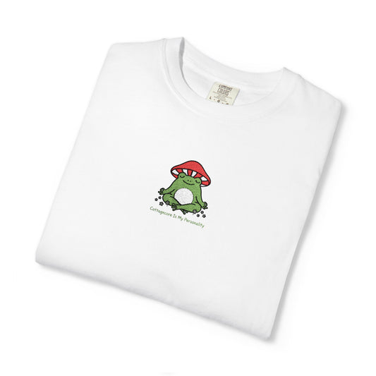 Close-up of a white Comfort Colors® shirt featuring an embroidered green frog meditating under a red mushroom with tiny flowers and the saying "Cottagecore Is My Personality."



