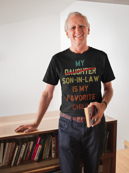Older man wearing a “My Son-in-Law Is My Favorite Child” funny family t-shirt with colorful text and bold strike-out effect, holding a book and smiling in a cozy home library setting—perfect for Father’s Day, dad gifts, family humor, and son-in-law appreciation tees.